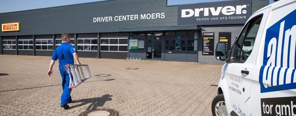 AlmTor-Mitarbeiter läuft von einem parkenden Firmenfahrzeug zu einer Werkstatt mit mehreren Garagen, demonstriert unseren engagierten Vor-Ort-Service und Kundennähe.