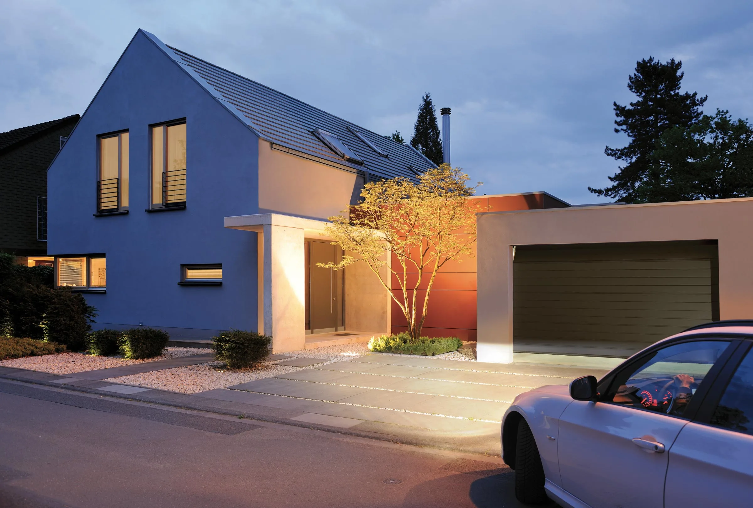 Modernes Haus in einer Wohnsiedlung am Abend mit sich öffnender Garage und beleuchtetem Innenraum, symbolisiert ein herzliches Willkommen zu Hause. Tür Installation inklusive einbau